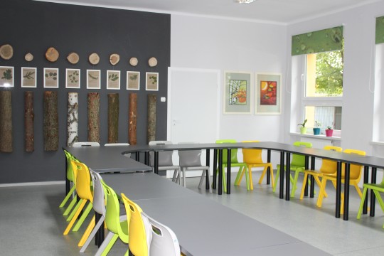 Na pierwszym planie stoły ustawione w kształcie litery U. Wokół rozstawione plastikowe, kolorowe krzesła. Po prawej stronie okna z roletami. Na tylnej ścianie dekoracje: obrazy, kawałki drewna.