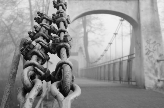 Czarno-białe zdjęcie zabytkowego mostu wiszącego. Na pierwszym planie łańcuch.