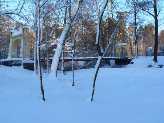 Na pierwszym planie śnieg zalegający na gruncie. Drzewa pozbawione liści. Na drugim planie most wiszący również pokryty śniegiem.