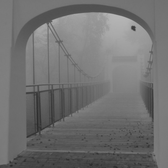 Czarno-białe zdjęcie mostu. Murowany łuk mostu na pierwszym planie, na drugim drewniane deski, liny i balustrada. Obraz we mgle.
