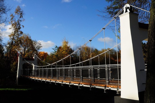 Na pierwszym planie most wiszący, po prawej murowany łuk, w środku liny i balustrada . Po prawej i po lewej stronie drzewa.
