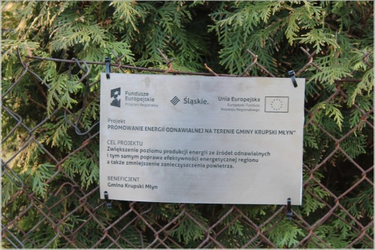 Na pierwszym planie tabliczka informacyjna, przymocowana do ogrodzenia z siatki. Na drugim planie drzewa iglaste.