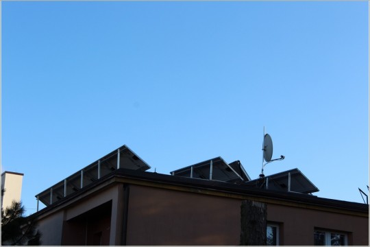 Na pierwszym planie dach budynku, na nim umiejscowione panele fotowoltaiczne. Powyżej błękitne, bezchmurne niebo.