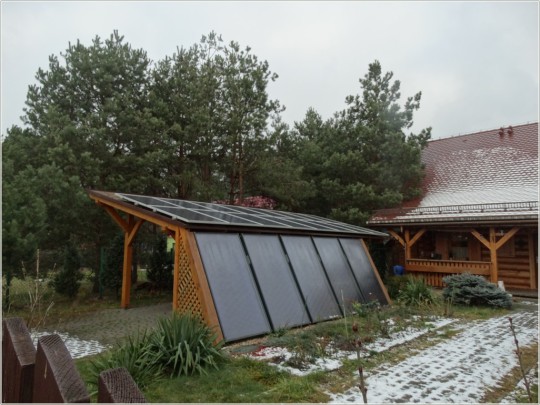 Widok na altanę. Na dachu i jednej ścianie altany panele fotowoltaiczne. Widoczny śnieg na trawniku. Wokół roślinność.