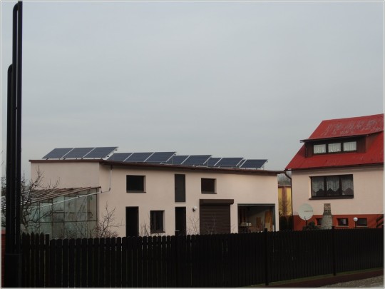 Widok na budynek gospodarczy, na dachu panele fotowoltaiczne. Po prawej stronie dom. Po lewej stronie w tle szklarnia przydomowa.