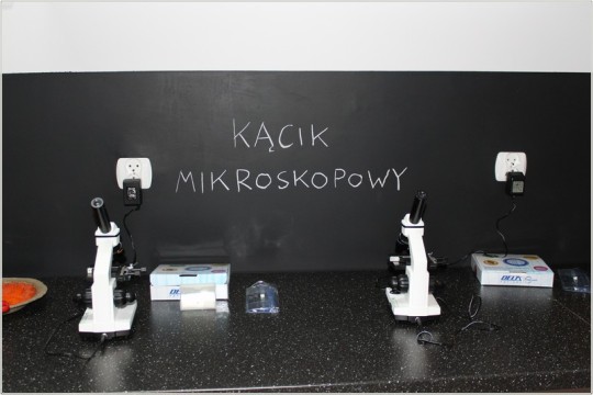 Białe mikroskopy na czarnym blacie. W tle czarna ściana z napisem "Kącik mikroskopowy".