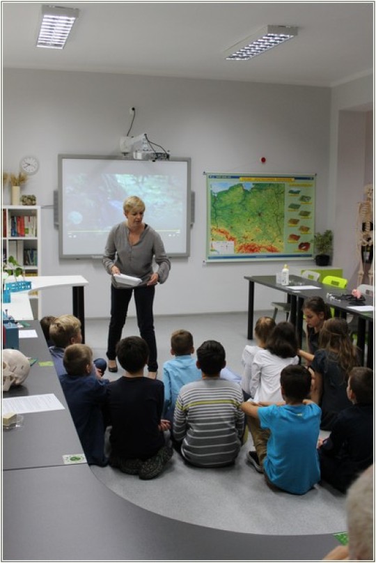 Na pierwszym planie grupa dzieci siedzi na macie, na środku klasy, są skierowani plecami. Na drugim planie kobieta, za kobietą tablica interaktywna. Dookoła stoły. Po prawej stronie w tle, mapa Polski.