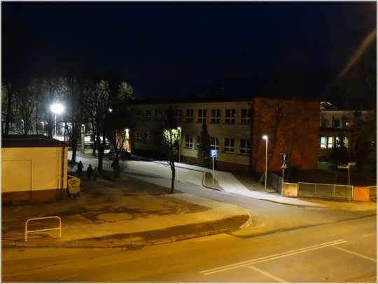 Widok z wysokości na drogę asfaltową, oświetloną lampami ledowymi. W tle budynek szkoły.