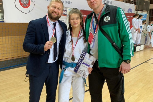 W środku dziewczyna, Mistrzyni Polski w Judo w kimono, w ręce trzyma dyplom. Na szyi zawieszony złoty medal. Z lewej mężczyzna w garniturze. w ręce trzyma złoty medal. Z prawej mężczyzna w dresie.