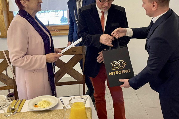 mężczyzna z prawej wręcza mężczyźnie z lewej prezent w czarnej torebce ze złotym napisem. z prawej stoi kobieta i trzyma teczkę. W tle mężczyzna w garniturze stoi bokiem, patrzy w lewą stronę