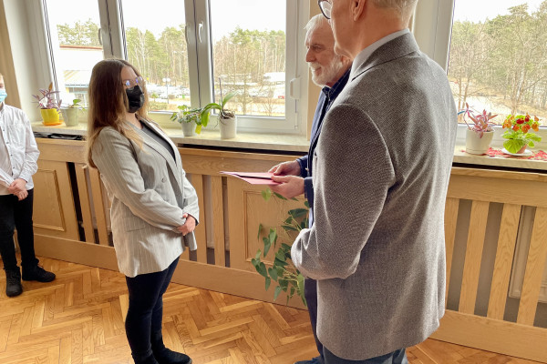 Z prawej 2 mężczyzn - Wójt i Przewodniczący Rady Gminy  wręczają młodej dziewczynie akt stypendialny w bordowej teczce. Dziewczyna zakrywa nos i usta czarną maseczką ochronną. W tle okno z widokiem na drzewa. Pod oknem zabudowa kaloryfera