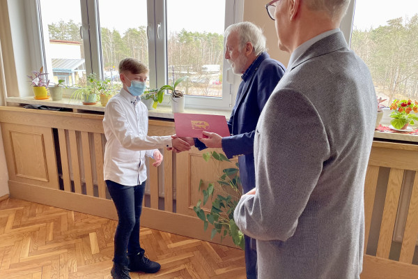 Z prawej 2 mężczyzn - Wójt i Przewodniczący Rady Gminy  wręczają młodemu chłopcu akt stypendialny w bordowej teczce. Chłopiec zakrywa nos i usta maseczką ochronną.  Wójt gratulując, ściska dłoń chłopca. W tle okno z widokiem na drzewa. Pod oknem zabudowa kaloryfera