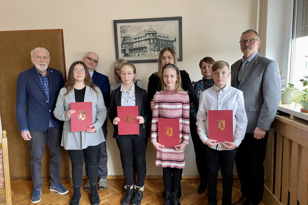 Na pierwszym planie grupa młodzieży trzymająca w rękach bordowe teczki ze złotym zdobieniem. Za nimi, na tle ściany z obrazem, stoją trzech mężczyzn i trzy kobiety.
