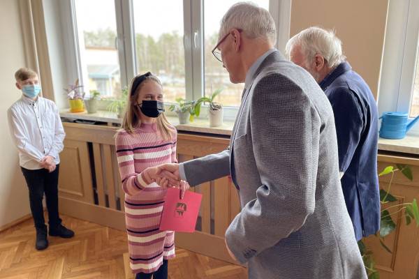 Z prawej 2 mężczyzn - Wójt i Przewodniczący Rady Gminy  wręczają młodej dziewczynie akt stypendialny w bordowej teczce. Dziewczyna zakrywa nos i usta czarną maseczką ochronną.  Przewodniczący Rady Gminy gratulując, ściska dłoń dziewczynki. W tle okno z widokiem na drzewa. Pod oknem zabudowa kaloryfera. W tle z lewej strony stoi chłopiec i przygląda się