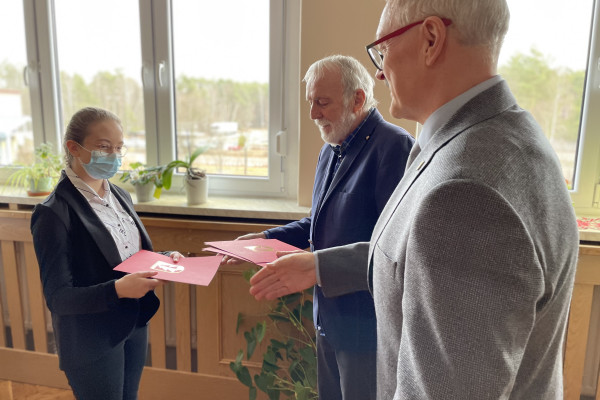 Z prawej 2 mężczyzn - Wójt i Przewodniczący Rady Gminy  wręczają młodej dziewczynie akt stypendialny w bordowej teczce. Dziewczyna zakrywa nos i usta  maseczką ochronną.  Przewodniczący Rady Gminy wyciąga dłoń chcąc pogratulować, dziewczynce. W tle okno z widokiem na drzewa. Pod oknem zabudowa kaloryfera.