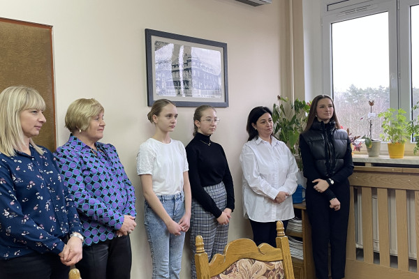 Na pierwszym planie stoi sześć kobiet , za nimi ściana. Po prawej stronie okno z widokiem na drzewa. Na parapecie stoją kwiaty.