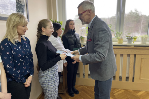 Na pierwszym planie stoją dwie kobiety i mężczyzna, który jednej z kobiet wręcza teczkę z aktem stypendialnym. Na drugim planie dwie kobiety. Za nimi okno z widokiem na drzewa. Na parapecie stoją kwiaty.