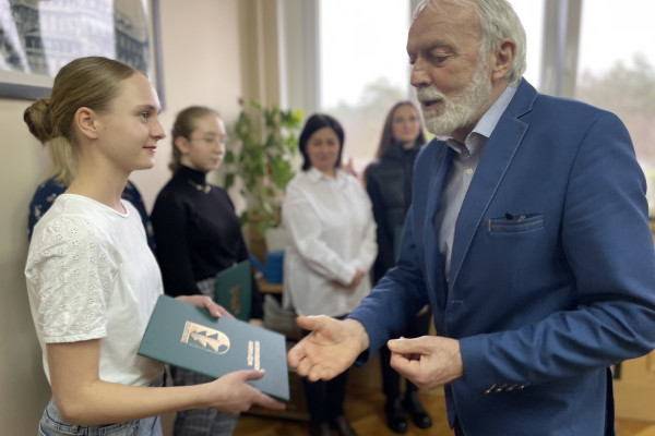 Na pierwszym planie stoją  kobiet i mężczyzna, który wręcza teczkę z aktem stypendialnym. Na drugim planie stoją dwie kobiety. Za nimi ściana a na niej wisi obraz