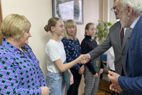 Na pierwszym planie stoją dwie kobiety i dwóch mężczyzn. Jeden z nich gratulując trzyma rękę kobiety. Na drugim planie dwie kobiety. Za nimi okno z widokiem na drzewa. Na parapecie stoją kwiaty.