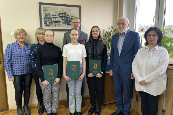 Na zdjęciu stoi grupa ludzi: trzy kobiety, dwóch mężczyzn  i trzy nastolatki trzymające w rękach teczki z logo gminy. Za ich plecami ściana z obrazem a po prawej stronie okno