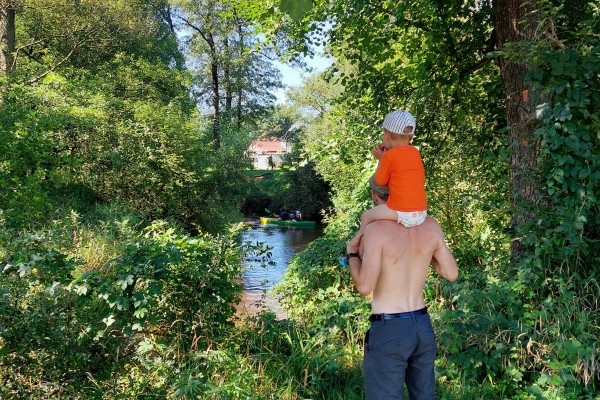 Z prawej tyłem stoi mężczyzna a na ramionach siedzi dziecko. Patrzą przed siebie na rzekę, po której płyną kajaki. Wokół zieleń lasu a w oddali budynek