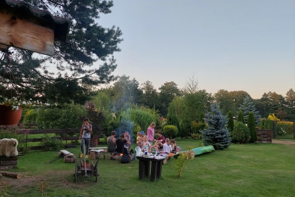W centralnej części fotografii przy ognisku siedzi grupa ludzi. W tle drzewa. U góry i z lewej zwisające gałęzie choinki.